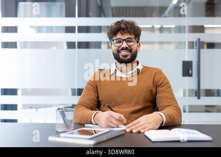 Young man with beard and glasses sitting at a modern office desk, smiling at the camera while taking notes in a notebook, symbolizing productivity and success Stock Photo