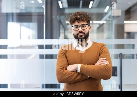 Young male business professional with glasses and beard stands arms crossed in a modern office, projecting confidence, leadership and career-focused professionalism Stock Photo