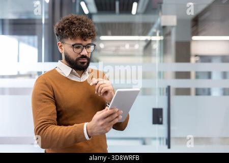 Young businessman with curly hair and glasses stands in a bright modern office, focused on a digital tablet while reviewing data, messaging, and planning strategy Stock Photo