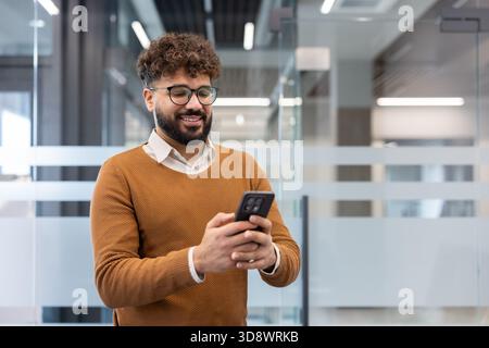 Young bearded man with curly hair smiling as he uses his smartphone in a modern office, conveying connectivity, mobile technology and professional, relaxed communication Stock Photo