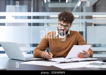 Young man with curly hair and glasses focusing on writing notes and reviewing documents, sitting at his desk with a laptop and papers in a modern office environment Stock Photo