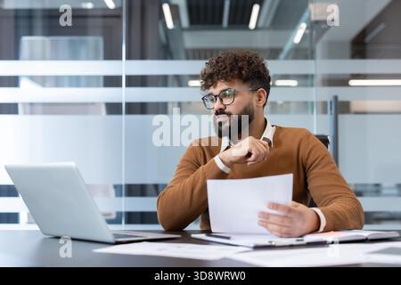 Young professional businessman with beard and glasses focused on laptop and paperwork at a modern office desk, analyzing reports, charts and data for strategic planning and projects Stock Photo