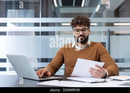 Young man with curly hair and glasses concentrating on his work, typing on a laptop and reviewing reports at an office desk, illustrating productivity and dedication Stock Photo