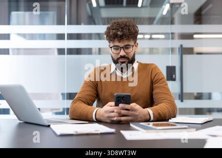 Young male professional concentrating on his cell phone while sitting at a desk with a laptop and documents in a contemporary office, connecting remotely Stock Photo