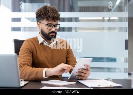 Young businessman with curly hair and glasses working on a digital tablet at his desk, analyzing important financial data and information in a modern business office environment Stock Photo