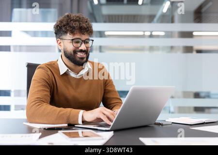 Young smiling businessman with glasses and beard working efficiently on his laptop at a modern corporate office desk, typing and managing his digital tasks with positive engagement and focus Stock Photo