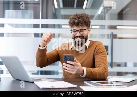 Enthusiastic young man with glasses celebrating success by clenching his fist and looking at his smartphone in a modern office, feeling excited and winning Stock Photo