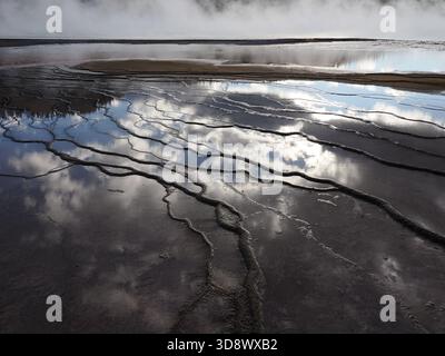 Detailed shot of blue sky and clouds reflected in the shallow, terraced water channels of a geothermal area, surrounded by dark bacterial mats. Stock Photo