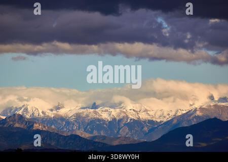 Snow-Capped Mountain Range Under Dramatic Clouds With Rolling Foreground Hills and Blue Sky Stock Photo