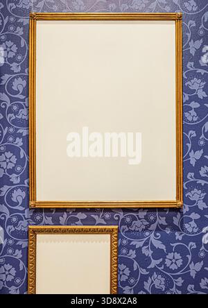 Two blank golden picture frames hanging on a dark blue wall with vintage floral pattern wallpaper. Stock Photo