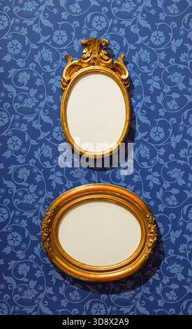 Two antique oval gold frames hanging on a blue wall with floral damask wallpaper. Stock Photo
