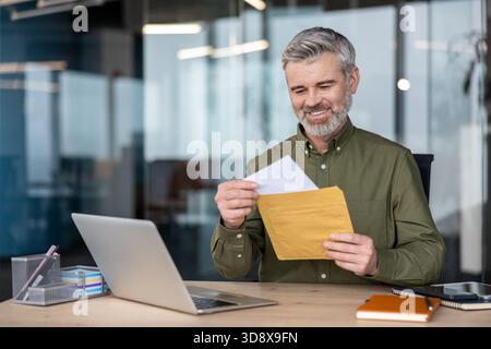 Mature cheerful businessman sitting at his office desk, happily opening a physical mail envelope with important documents, an old-school alternative to digital correspondence, while using a laptop Stock Photo