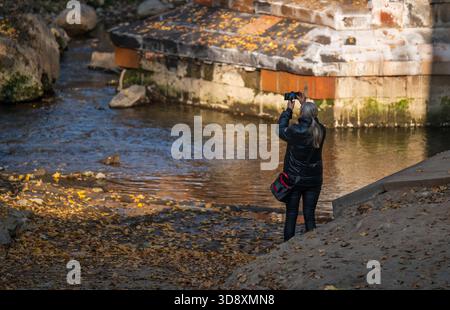 Yerevan Armenia 10.25.2025. A woman is taking a picture of a river with a camera. The water is calm and the leaves are falling from the trees Stock Photo