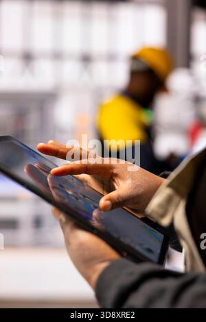 Industry 4.0 engineer programs automated assembly line and robot arm using CAD software on tablet. African American automation specialist employs digital interface to control advanced manufacturing. Stock Photo