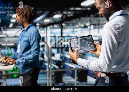 African american data center staff reviews server rigs on industrial platform, using a tablet for data management and innovation. Engineer coordinating infrastructure tasks for machine learning. Stock Photo