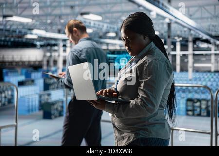 Server room coworkers performing maintenance on server racks using software on laptop. Teamworking data center technicians upgrading blade server components using notebook Stock Photo