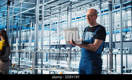 Elderly engineer uses laptop in smart factory to test automated systems, ensuring reliability. Experienced technician uses notebook to identify bottlenecks and optimize equipment, camera A Stock Photo