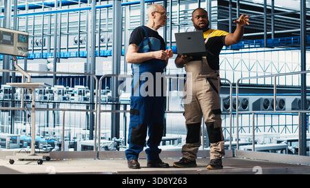 Senior engineer talking with intern in smart factory, collecting and analyzing data. Experienced technician helping trainee use tracking software in industrial plant equipped with sensors, camera A Stock Photo
