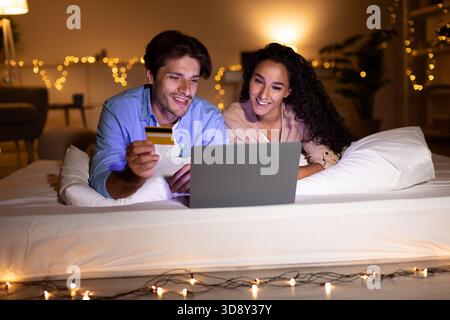 Happy Couple Enjoys Online Shopping Together at Night With Laptop on Mattress Stock Photo