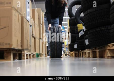 A low angle shot of a woman rolling or installing a new car tire down an aisle between stacks of boxes and tires in a store. High quality photo Stock Photo