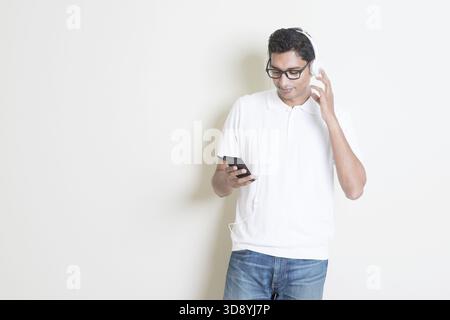 Smartphone social media concept. Indian guy using mobile phone. Asian man relaxed and listening music with headset, standing on plain background. Hand Stock Photo