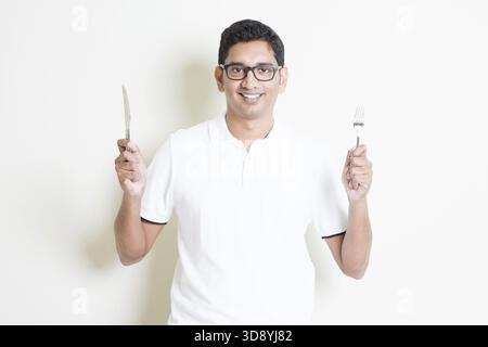 Dining concept. Indian guy holding cutlery fork and knife on hand, hungry for food. Asian man standing on plain background with shadow and copy space. Stock Photo