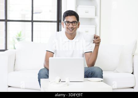 Young Indian guy working from home with laptop computer. Asian man using internet indoor, enjoying cup of tea, relaxed and sitting on sofa. Handsome m Stock Photo