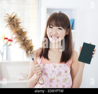 Happy Asian housewife with apron housecleaning, hand holding duster and cleaning sponge. Young woman indoors living lifestyle at home Stock Photo