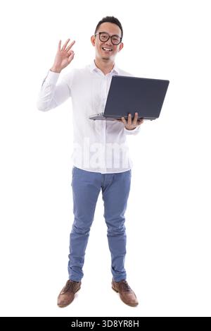 Full length of happy Asian businessman using laptop showing okay hand sign standing isolated over white background Stock Photo