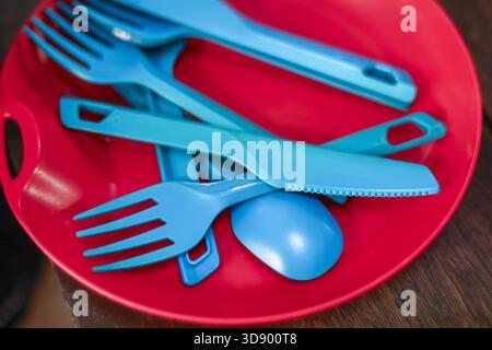 Close up of blue plastic cutlery including fork, spoon, and knife inside red bowl. These reusable utensils are ready for picnic or camping trip, simpl Stock Photo