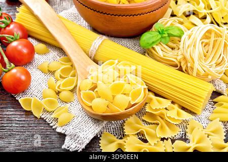 Pasta seashells in spoon, pipe rigate in a bowl, spaghetti, tagliatelle, penne, farfalle, trenette, tomatoes and parsley on burlap on dark wooden boar Stock Photo
