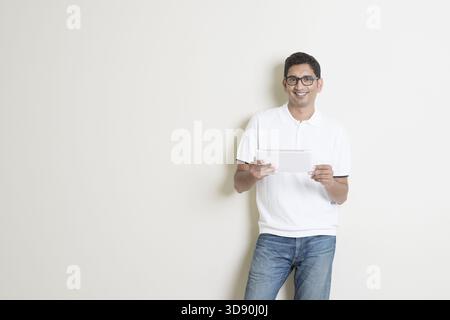 Portrait of handsome Indian guy using tablet pc, standing on plain background with shadow, copy space at side Stock Photo