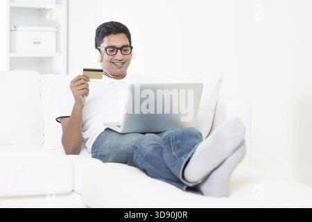 Indian guy making online credit card payment with laptop computer at home. Asian man internet shopping indoor relaxed and sitting on sofa. Handsome ma Stock Photo