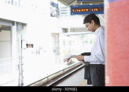 Candid Asian Indian businessman waiting at public train station, standing and reading on newspaper Stock Photo