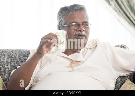 Elderly healthcare concept. Old Indian man drinking a glass water, sitting on sofa at home. Retired senior people living lifestyle Stock Photo
