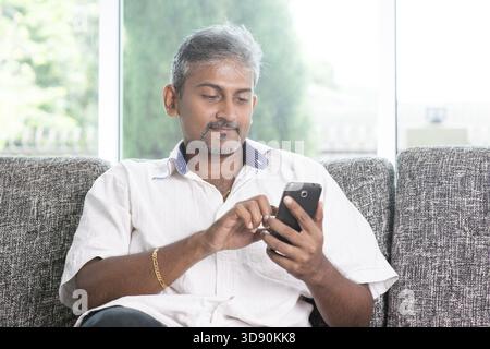 Smartphone social media concept. Indian mature guy using mobile phone. Asian man relaxed and sitting on sofa indoor Stock Photo