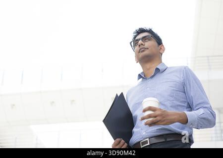 Young Asian Indian businessman holding a cup of coffee looking away, India male business man, real modern office building as background Stock Photo