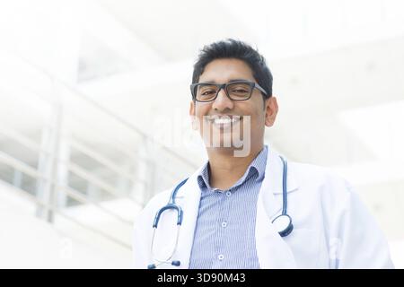 Portrait of a smiling Asian Indian male medical doctor in uniform, hospital building at background Stock Photo