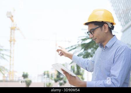 Asian Indian engineer looking at digital computer tablet, pointing to the construction site, inspecting the progress of project Stock Photo