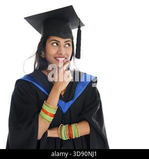 Happy Indian graduate student in graduation gown and cap thinking and smiling. Portrait of beautiful Asian female model standing isolated on white bac Stock Photo