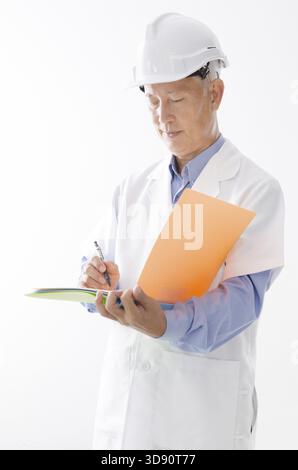 Portrait of old Asian contractor in uniform with hard hat writing, standing isolated on white background Stock Photo