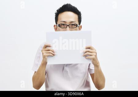 Portrait of sad Asian man hand holding white blank paper card covering his eyes, standing isolated on plain background Stock Photo