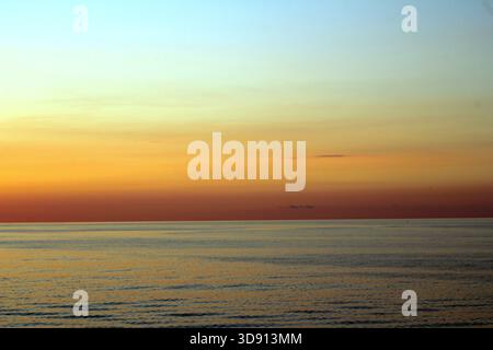 Calm Ocean Horizon at Dusk with Golden Sky. A serene, wide-angle view of the ocean horizon just after sunset. The sky transitions from pale blue Stock Photo