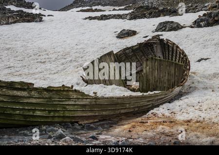 Remains of an ancient boat, on the rocky shore of one of the Antarctic islands. Peninsula. Snow, rocky hills in background. Stock Photo