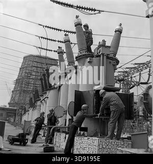KRAMATORSK, USSR - 1976: A team of Soviet electrical workers performs maintenance on high-voltage oil circuit breakers at the Kramatorsk Combined Heat and Power Plant substation. Mechanics in safety helmets climb the ceramic bushings while others adjust the control mechanism at ground level. In the background, a massive hyperboloid cooling tower looms over the site. A scene of collective industrial labor ensuring the energy stability of the peaceful Donbass region during the Soviet era. Stock Photo