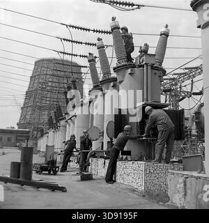 KRAMATORSK, USSR - 1976: A team of Soviet electrical workers performs maintenance on high-voltage oil circuit breakers at the Kramatorsk Combined Heat and Power Plant substation. Mechanics in safety helmets climb the ceramic bushings while others adjust the control mechanism at ground level. In the background, a massive hyperboloid cooling tower looms over the site. A scene of collective industrial labor ensuring the energy stability of the peaceful Donbass region during the Soviet era. Stock Photo