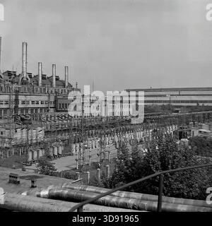 KRAMATORSK, USSR - 1976: General view of the Kramatorsk Combined Heat and Power Plant (CHP). The image features the main industrial building topped with a row of tall smokestacks and an extensive open-air electrical switchyard (substation) in the foreground. Numerous high-voltage circuit breakers, lattice steel structures, and insulated heating pipes fill the landscape. A panoramic snapshot of the massive energy infrastructure powering the heavy industry of the peaceful Donbass. Stock Photo