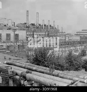 KRAMATORSK, USSR - 1976: General view of the Kramatorsk Combined Heat and Power Plant (CHP). The image features the main industrial building topped with a row of tall smokestacks and an extensive open-air electrical switchyard (substation) in the foreground. Numerous high-voltage circuit breakers, lattice steel structures, and insulated heating pipes fill the landscape. A panoramic snapshot of the massive energy infrastructure powering the heavy industry of the peaceful Donbass. Stock Photo