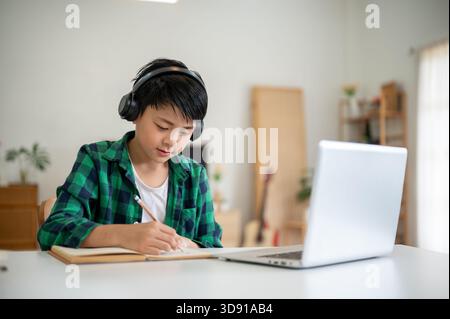 Asian kid elementary school boy wearing headphones writing with pencil in notebook with laptop aside. Online Study, Doing homework, internet Surfing. Stock Photo