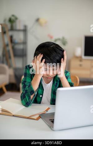 Asian kid elementary school boy looking tired while sitting with hands holding head in front of laptop. Online Study, Doing homework, Internet surfing Stock Photo